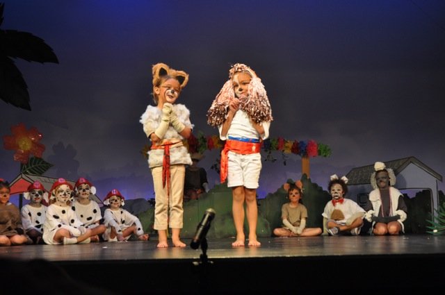 Two young performers in animal costumes sing at a microphone, with the cast seated behind them