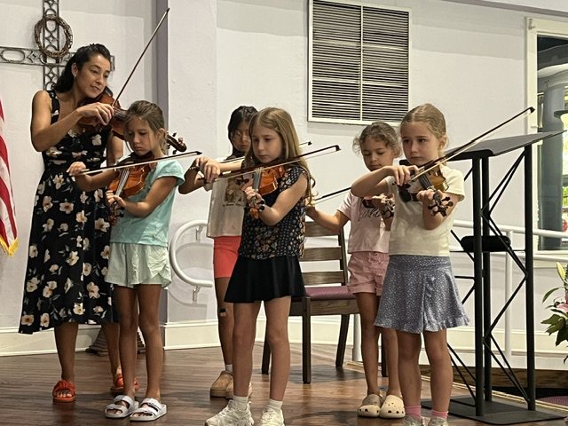 A teacher plays violin beside a row of young violinists